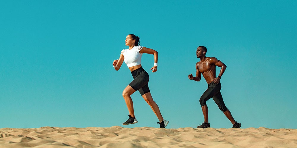 A male and female runner jogging across sand in front of blue sky.